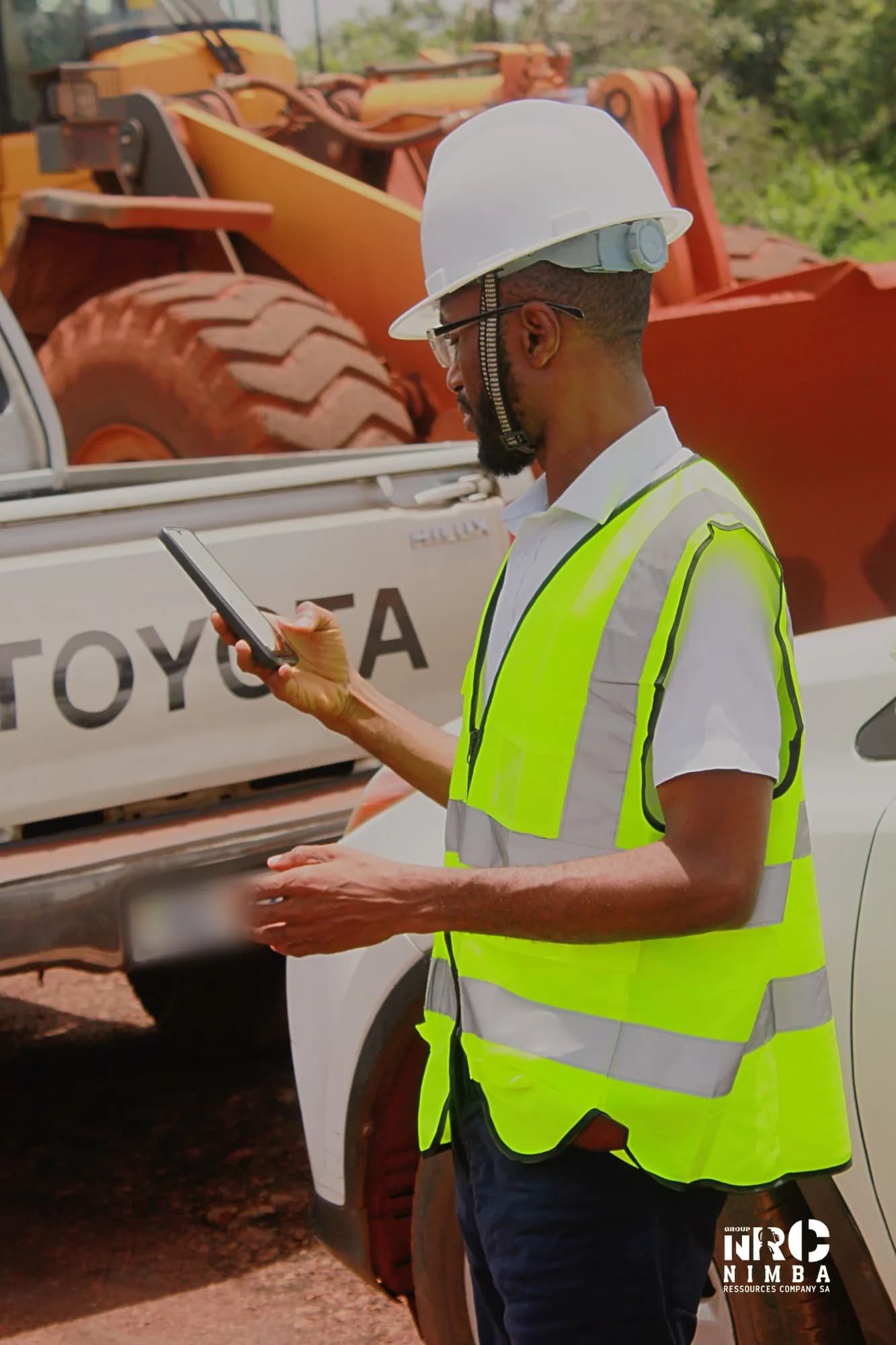 Personne en casque blanc et gilet de sécurité NRC consultant son téléphone devant un pick-up et un engin de chantier