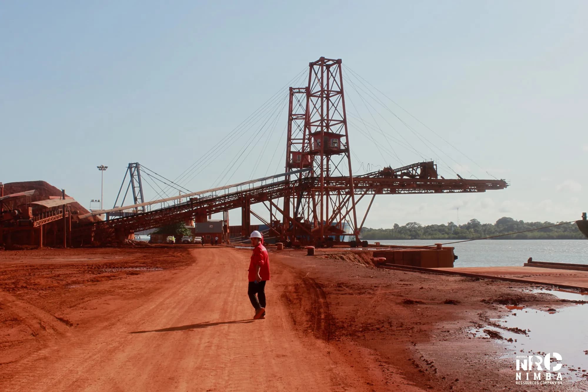 Personne en casque blanc marchant sur un quai portuaire devant une tour de chargement, au bord de l’eau